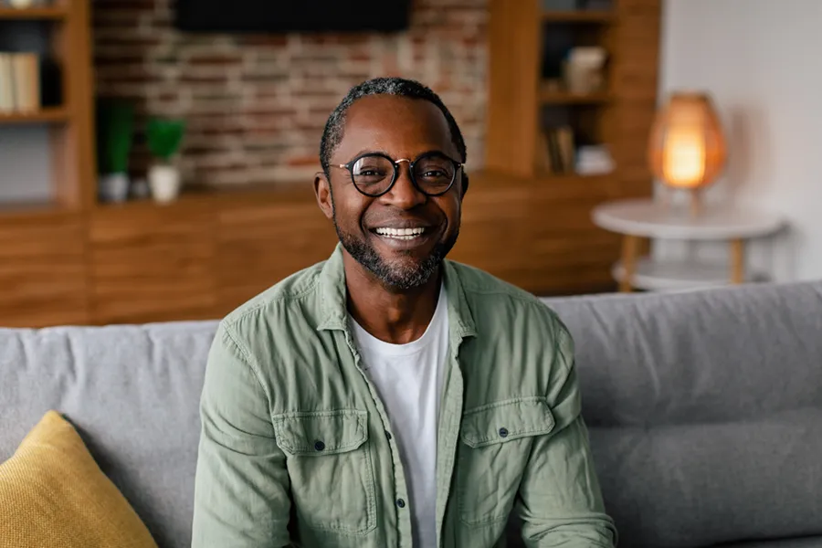 Andropause-Doctor A middle aged black man in a green button up shirt sits on his couch smiling, happy with his treatment for andropause and low testosterone from Lux Hormones & Health Wellness in San Diego.