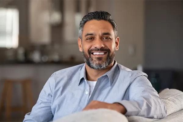 BioTE-Testosterone-Pellet-Clinic A mature South Asian man seated comfortably on a couch, smiling and looking directly at the camera satisfied with BioTE Testosterone Pellet Therapy offered by Lux Hormones & Health Wellness in San Diego.