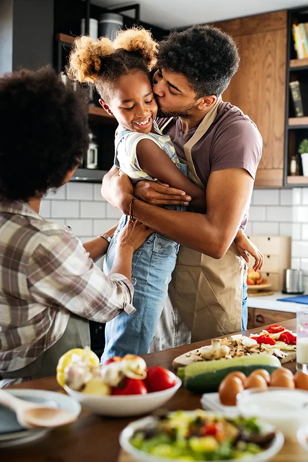 Gut-Health-Treatment A man holds his daughter and kisses her cheek as the family makes a healthy dinner in the kitchen. Get help for digestive issues and gut health from Lux Hormones & Health Wellness in San Diego.