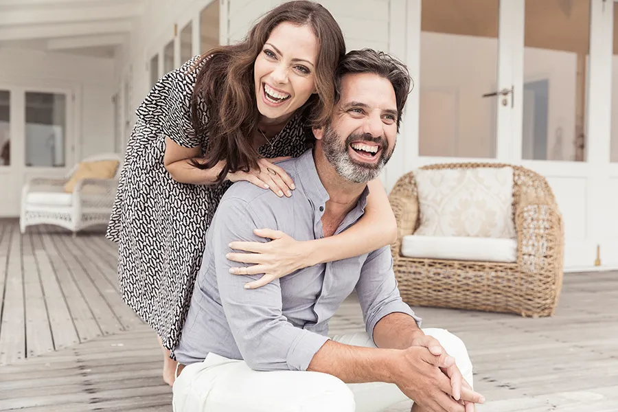 Health-and-Wellness-Services-Doctor A middle-aged couple on a wooden porch with wicker furniture; the woman is playfully hugging the man from behind. They are benefiting from the health and wellness services from Lux Hormones & Health Wellness in San Diego.