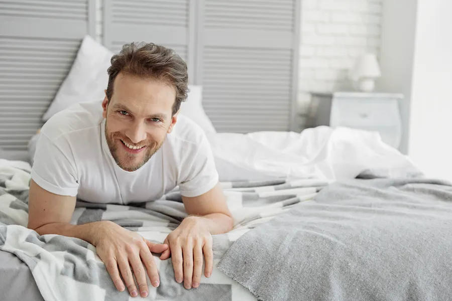 Low-Libido-in-Men-Doctor A dirty-blonde haired man lays on his stomach in bed smiling. Schedule treatment for low libido in men from Lux Hormones & Health Wellness in San Diego.