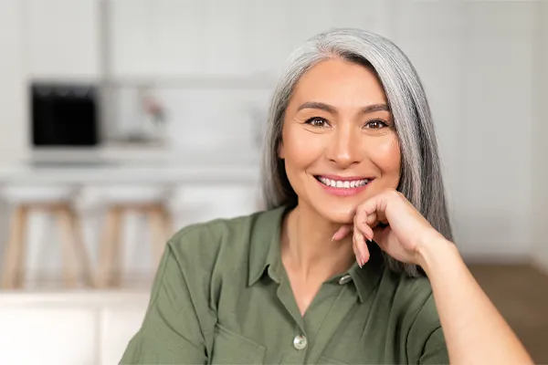 Menopause-Clinic An asian woman with long gray hair and a green shirt sitting and smiling on her couch, happy with her menopause care from [NAME} in San Diego.