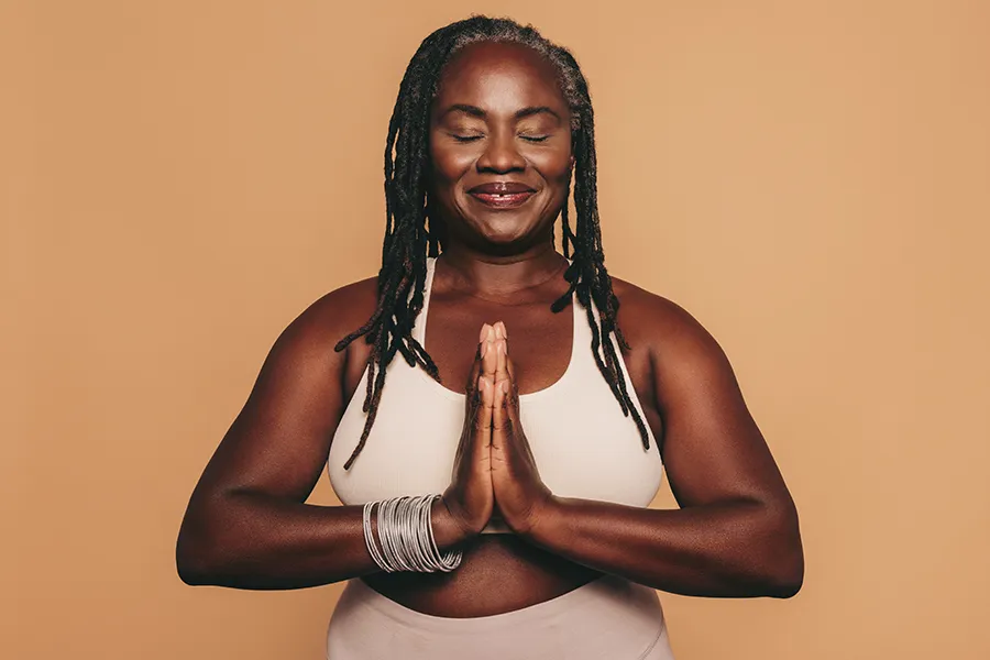 Obesity-Clinic An older African American woman confidently posing against a warm background smiling with hands in a prayer pose. Lux Hormones & Health Wellness treats obesity using functional medicine in San Diego.